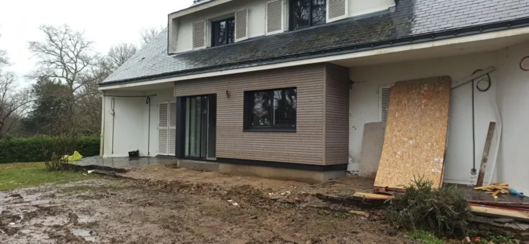 Une maison moderne à deux étages avec de grandes fenêtres, une cour partiellement boueuse et des matériaux de construction contre le mur.