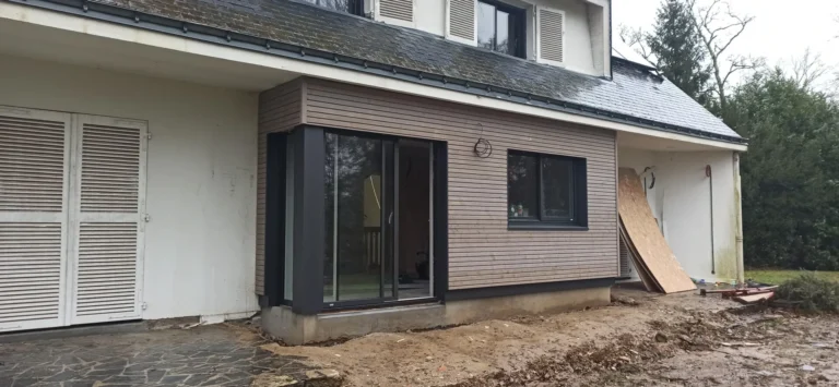Une maison avec une extension moderne en bois, une grande porte coulissante en verre et un espace extérieur non fini.