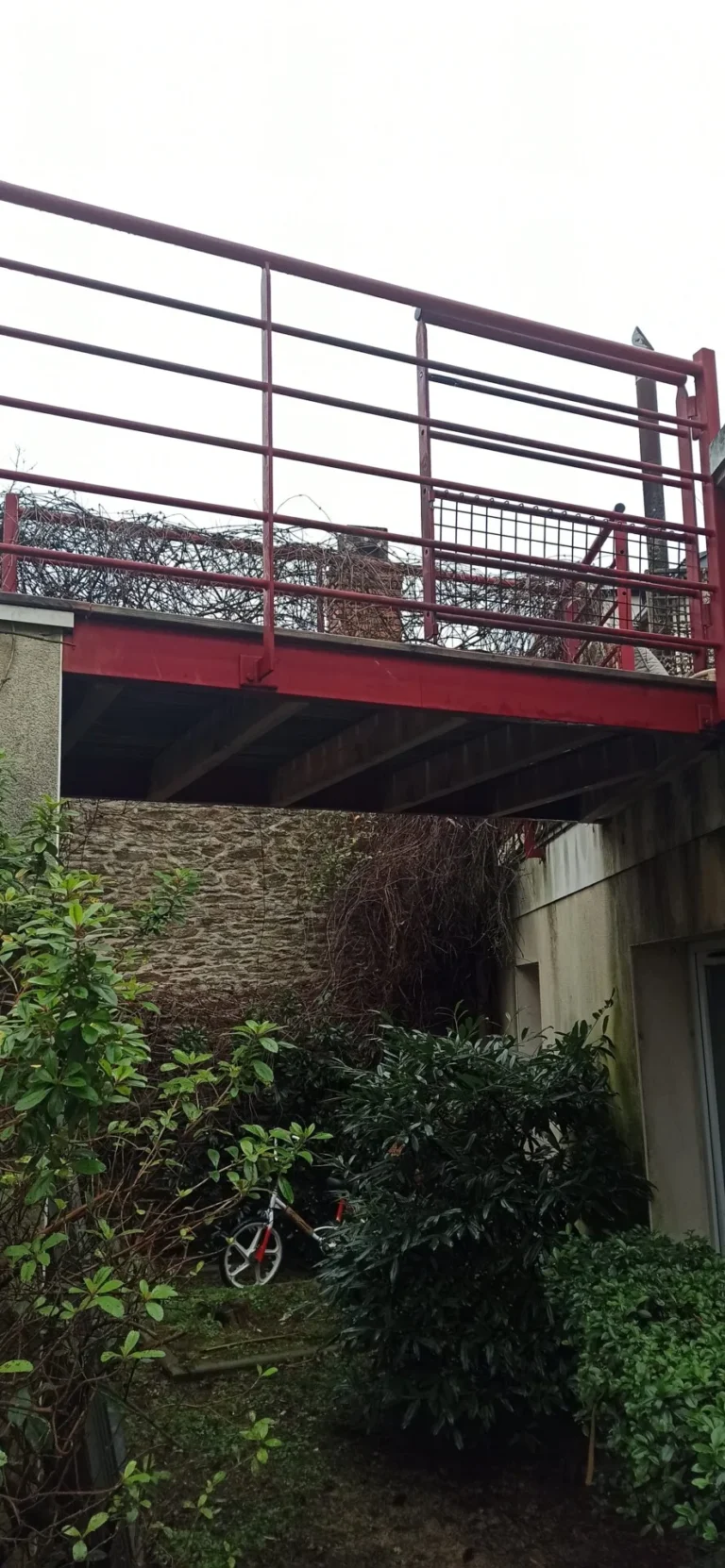 Un pont métallique rouge relie deux bâtiments au-dessus d'un jardin verdoyant avec une roue de vélo visible.