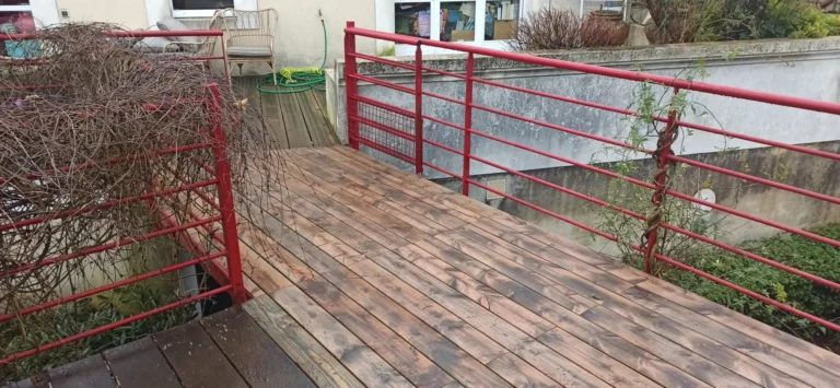Une passerelle en bois avec des balustrades rouges relie deux zones. Des plantes grimpantes ornent les balustrades et un tuyau d'arrosage apparaît en arrière-plan.