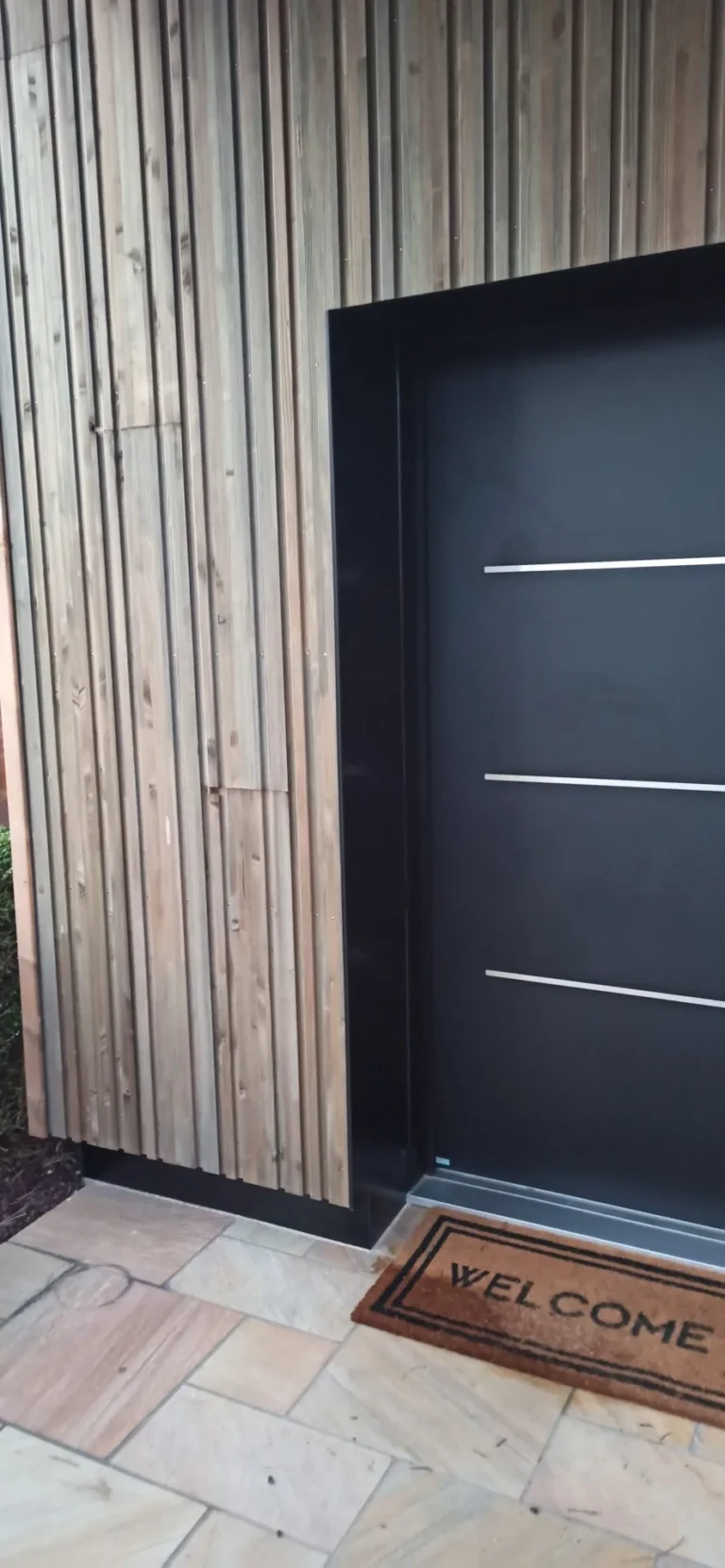 Une porte noire moderne aux lignes horizontales argentées est encastrée dans un mur en bois. Un paillasson « Bienvenue » est posé au sol devant.