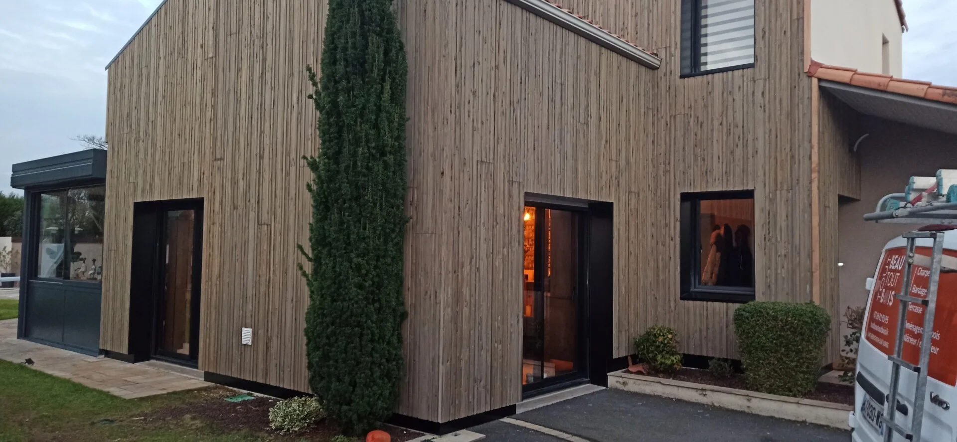 Maison moderne à deux étages avec extérieur en bois, avec un grand arbre à feuilles persistantes près de l'entrée, un intérieur éclairé par les fenêtres et un véhicule garé sur la droite.