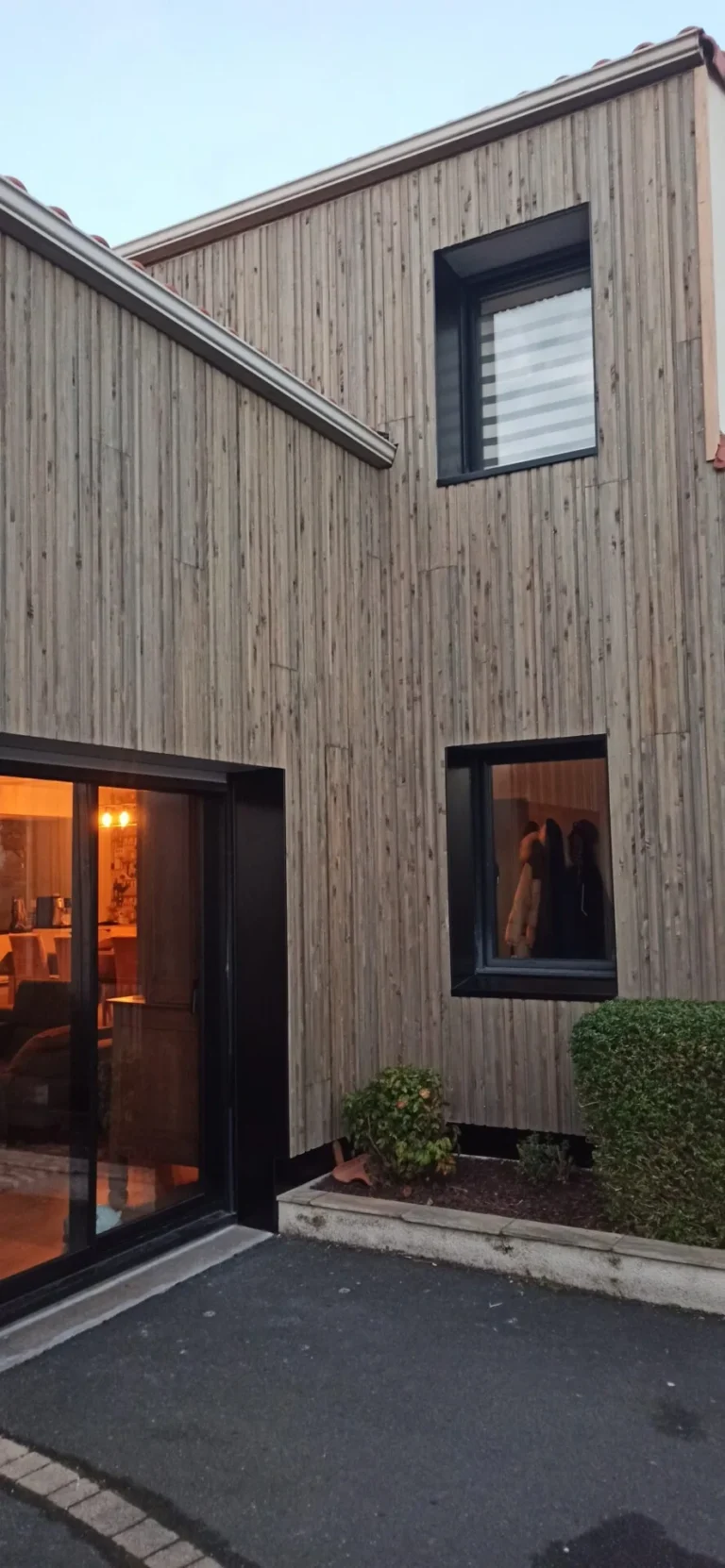 Façade d'une maison moderne en bois avec de grandes fenêtres, dont une éclairée de l'intérieur. De petits buissons bordent le trottoir adjacent.