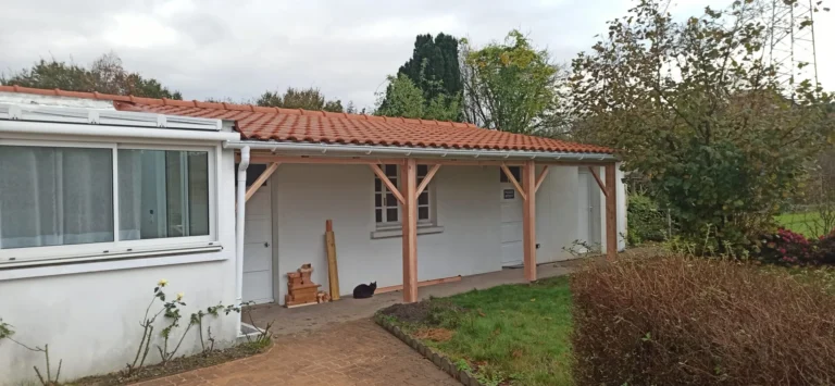 Une petite maison blanche au toit de tuiles rouges et au porche couvert. Des poutres en bois soutiennent le porche et un chat noir est assis près d'un objet en bois. Le jardin est entouré de buissons et d'arbres.