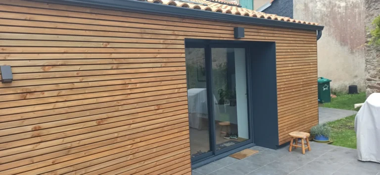 Mur extérieur en bois avec lames horizontales et grande porte coulissante en verre. Un carrelage gris mène à l'entrée. Un petit tabouret en bois se trouve près de la porte.