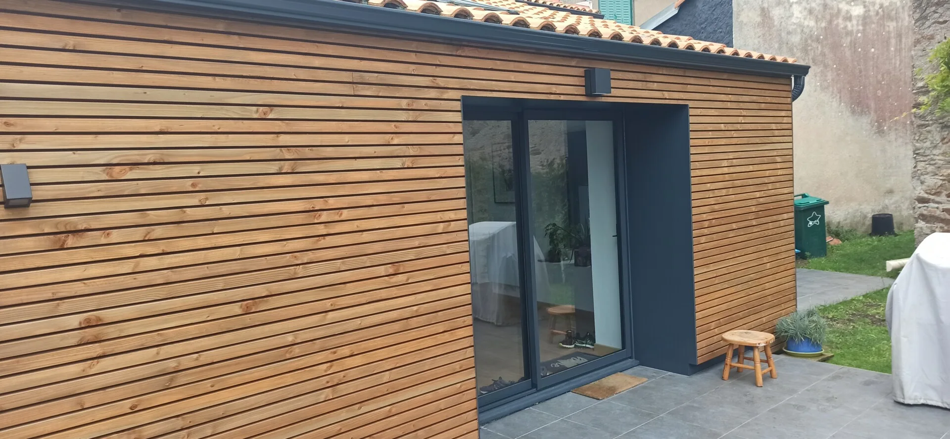 Mur extérieur en bois avec lames horizontales et grande porte coulissante en verre. Un carrelage gris mène à l'entrée. Un petit tabouret en bois se trouve près de la porte.