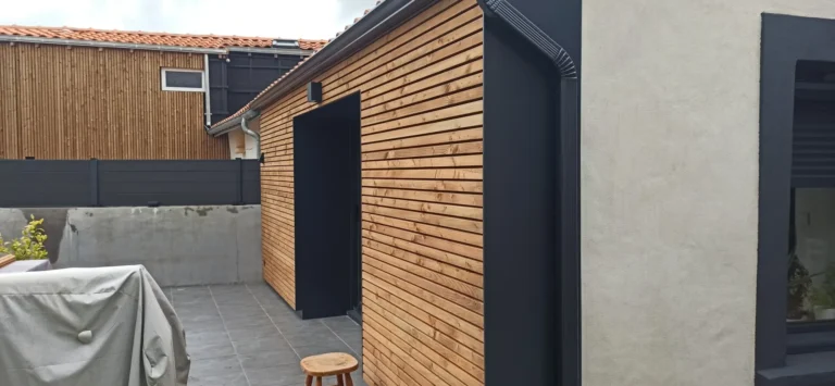 Façade de bâtiment lambrissée avec une petite fenêtre, adjacente à une terrasse carrelée. Un objet couvert et un tabouret en bois se trouvent sur la terrasse. À proximité se trouvent une clôture grise et une autre structure en bois.