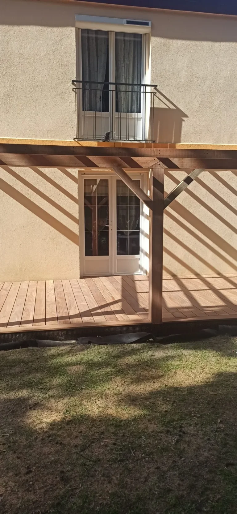 Terrasse en bois avec pergola devant une maison à deux étages avec portes-fenêtres et fenêtres à la française. Les ombres créent des motifs rayés sur la terrasse.