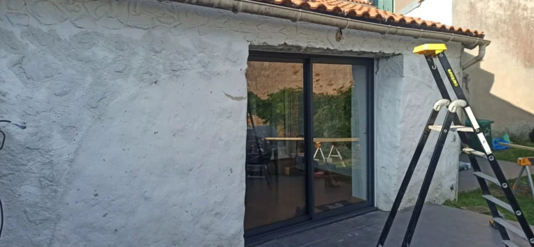 Un mur en stuc blanc avec de grandes portes coulissantes en verre. Un escabeau se trouve à droite. Les portes reflètent une scène extérieure et des lumières intérieures sont visibles à l'intérieur.