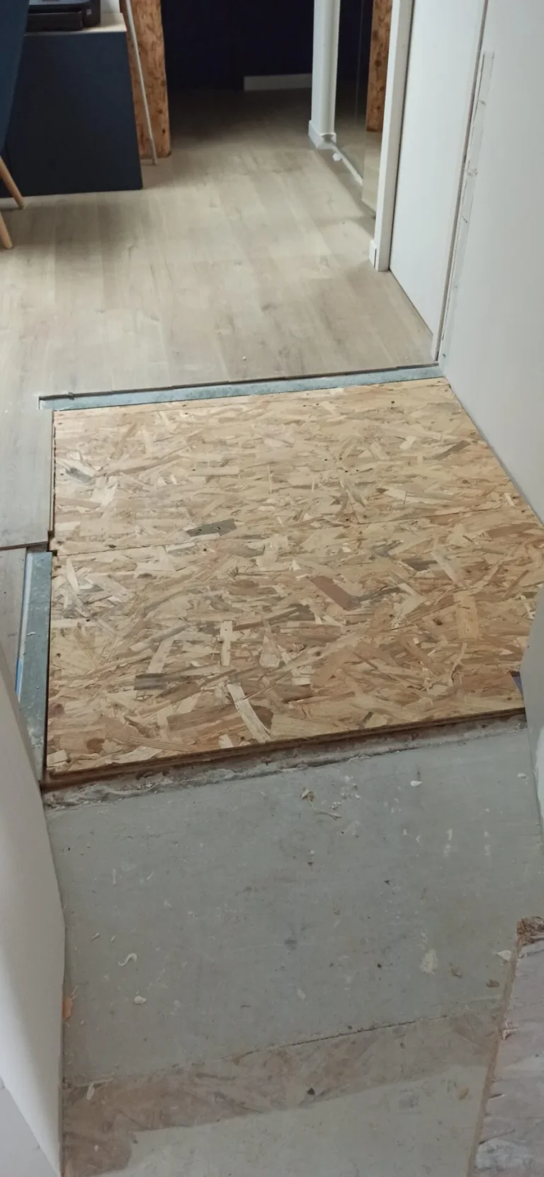 Un morceau de contreplaqué placé comme revêtement de fortune sur un espace dans le plancher entre deux pièces d'un bâtiment en cours de rénovation.