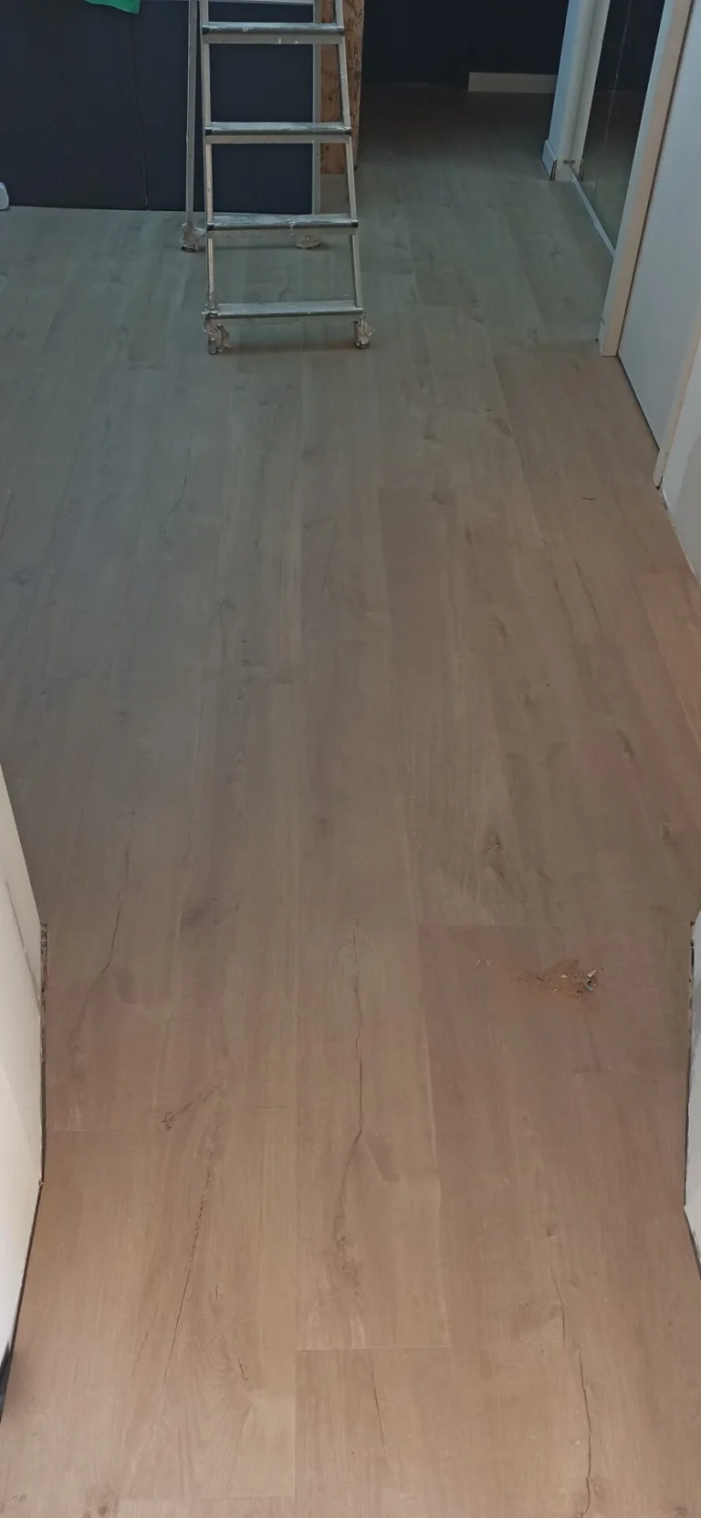 Plancher en bois légèrement endommagé au centre. Un petit escabeau se trouve au fond de la pièce, près d'un mur. La pièce semble être en cours de rénovation.