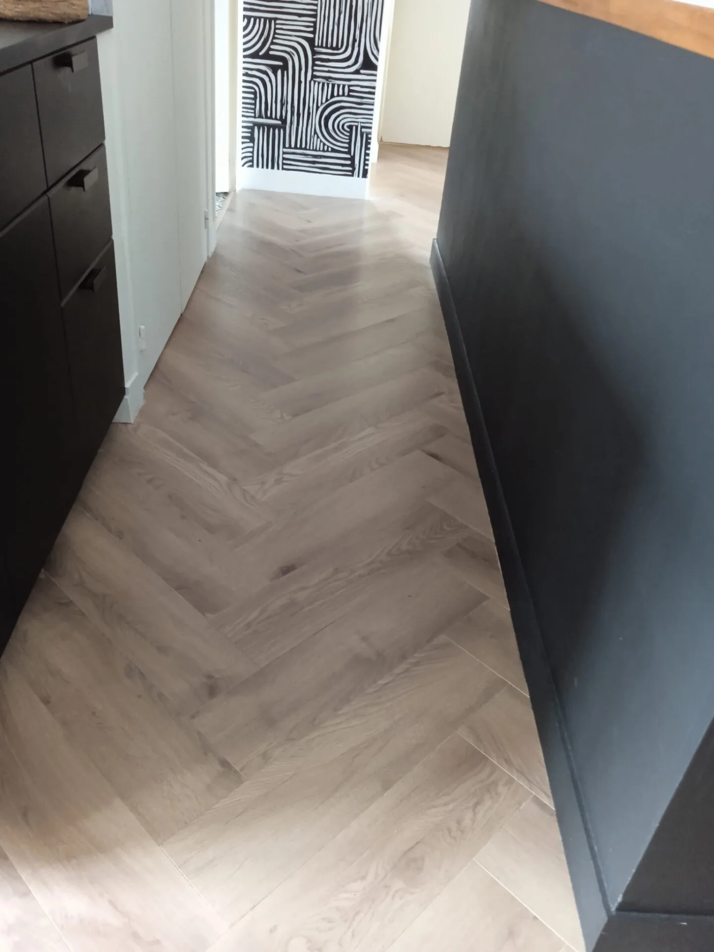 Un couloir avec un parquet à chevrons, des armoires sombres sur la gauche et un mur à motifs géométriques noir et blanc au bout.