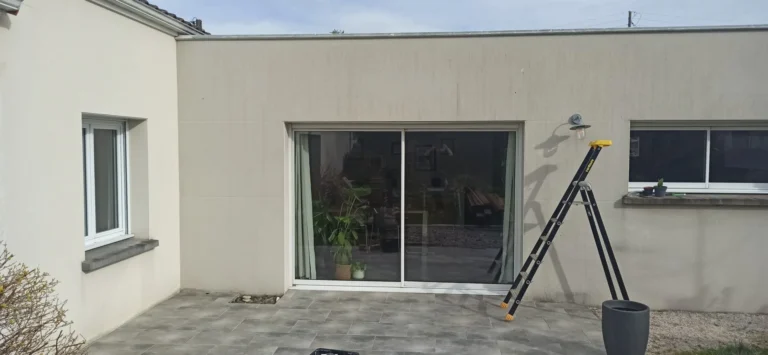 On voit un bâtiment beige avec une porte coulissante en verre. Une grande échelle est appuyée contre le mur, et une plante en pot est posée à côté de la porte.