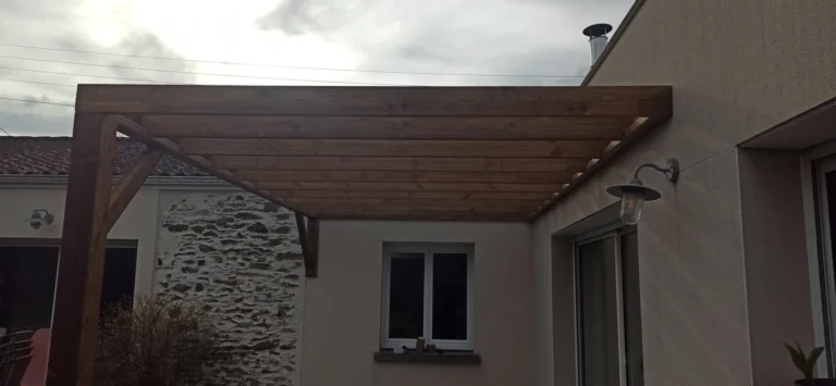 Pergola en bois adossée à un bâtiment, avec une applique murale de style vintage à proximité. Ciel couvert en arrière-plan.
